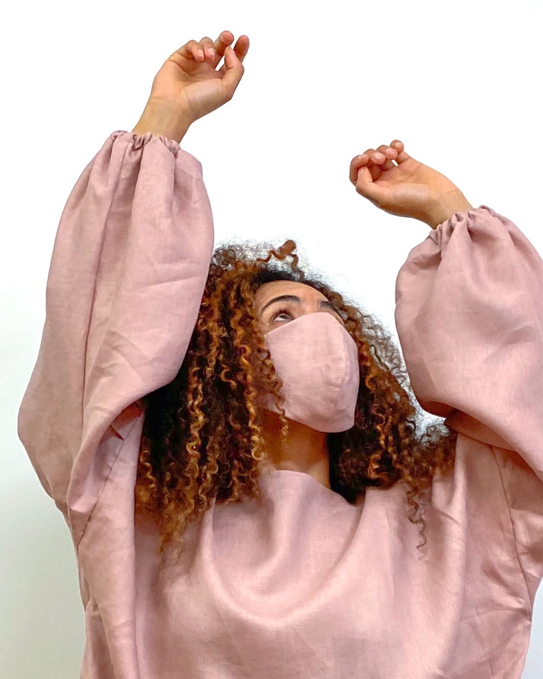 A woman wears a dusky pink linen dress with balloon sleeves, and a matching face mask. 