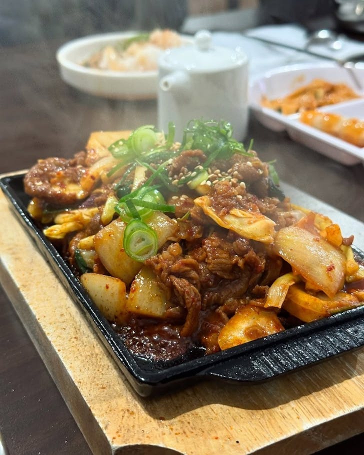 A steaming hot dish of stir-fried beef with onion, topped with spring onion and sesame.