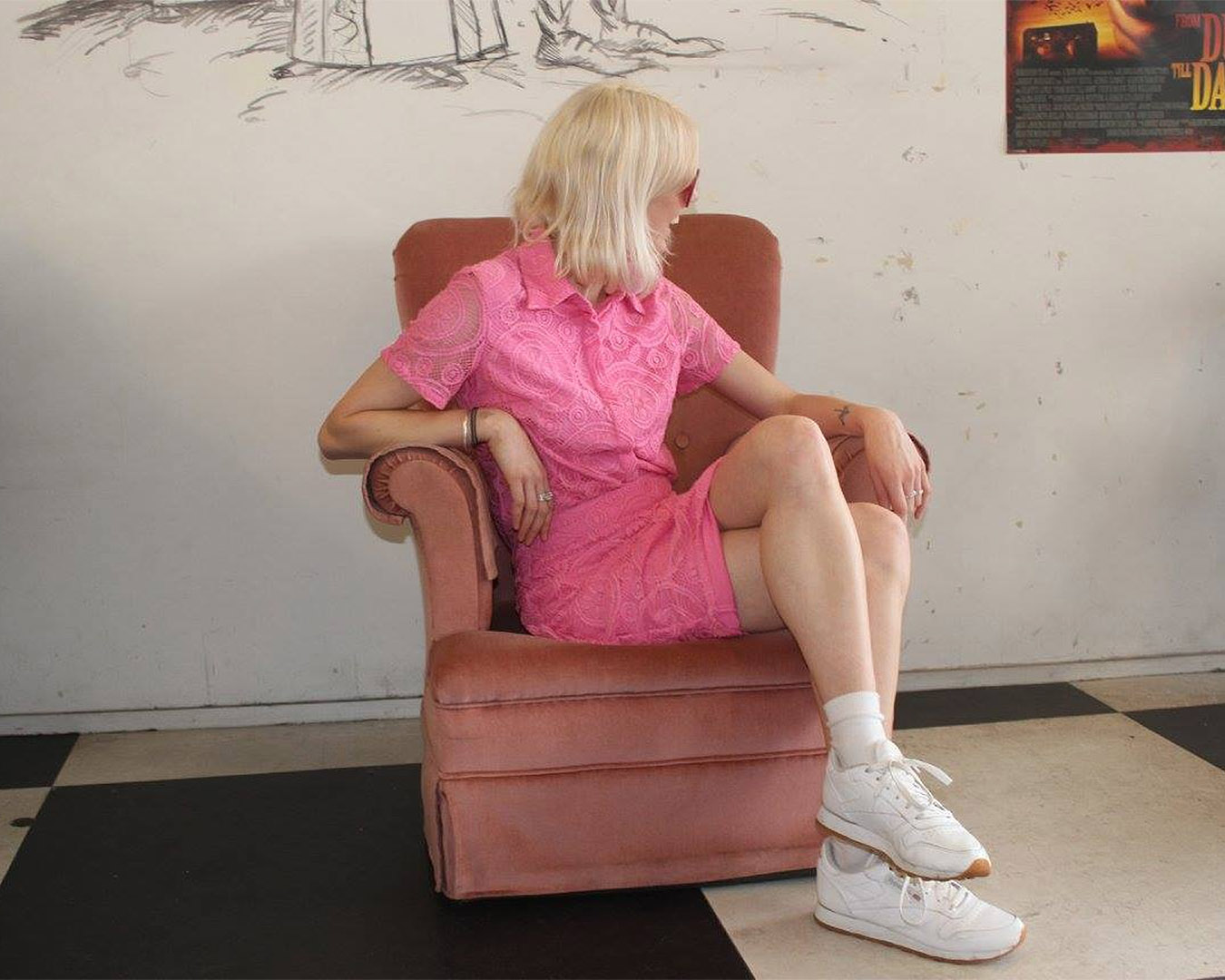 A woman sits on a dusky pink armchair wearing a light pink lacy dress as she looks off camera. 