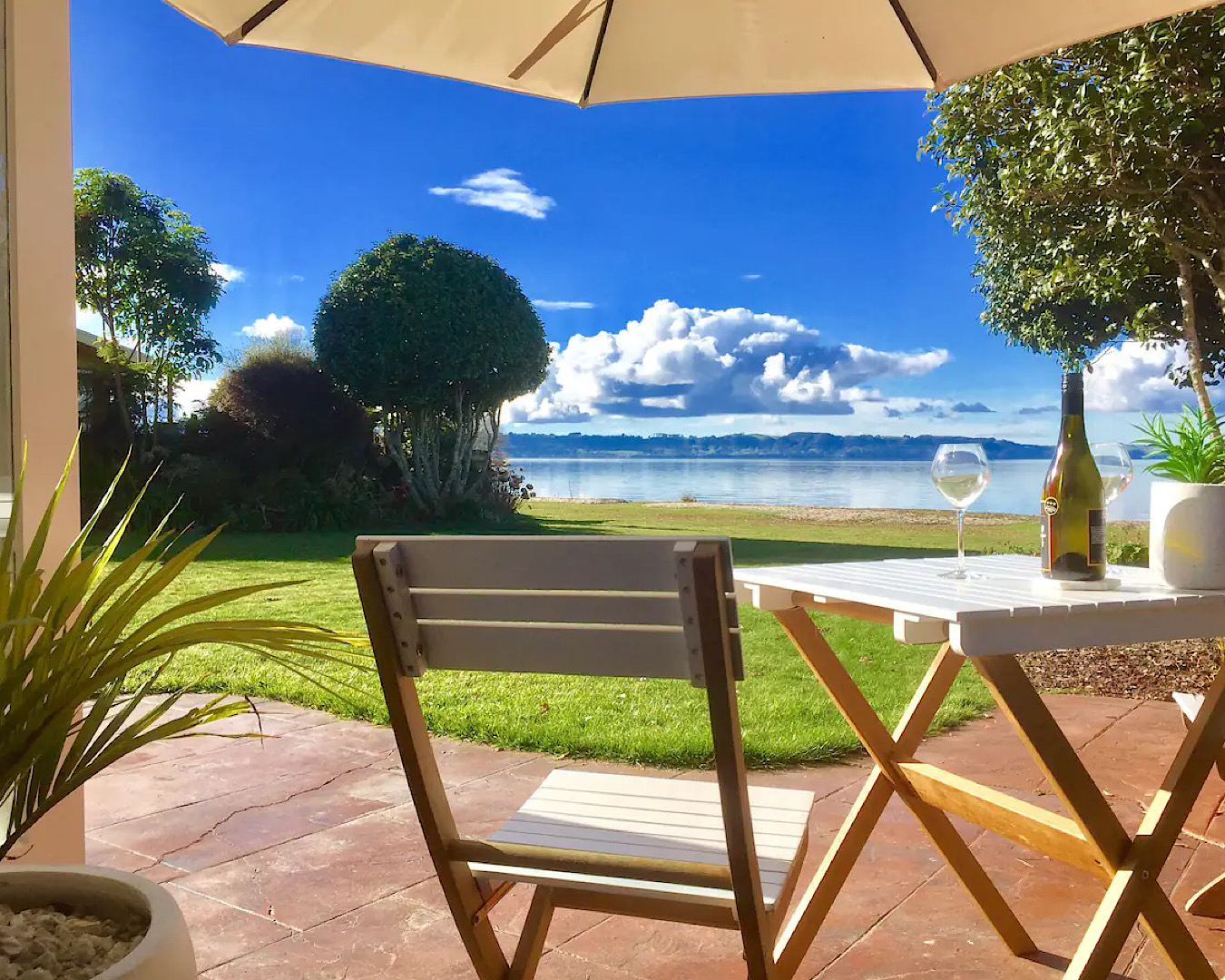 Blue skies, a lake view and two glasses of wine from a cute deck set up at Rotorua Airbnb, Parawai Bay Lakeside Retreat. 