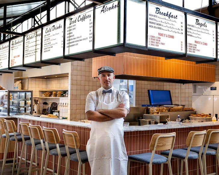 A man in a flat cap and bowtie stands with arms folded in front of the Pastrami and Rye American deli-style fit-out at Ponsonby Central. 