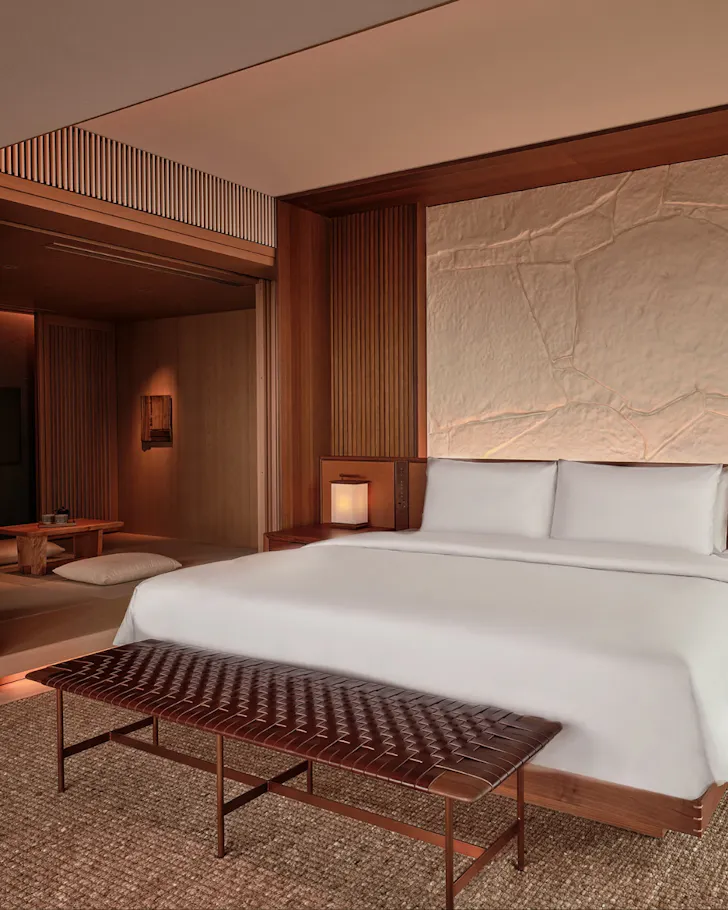A soothing suite with warm tones, a stone backdrop and crisp white bedding at Patina Osaka. 