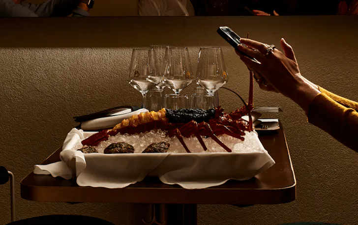A lobster on a table with a person taking photos on their phone. 
