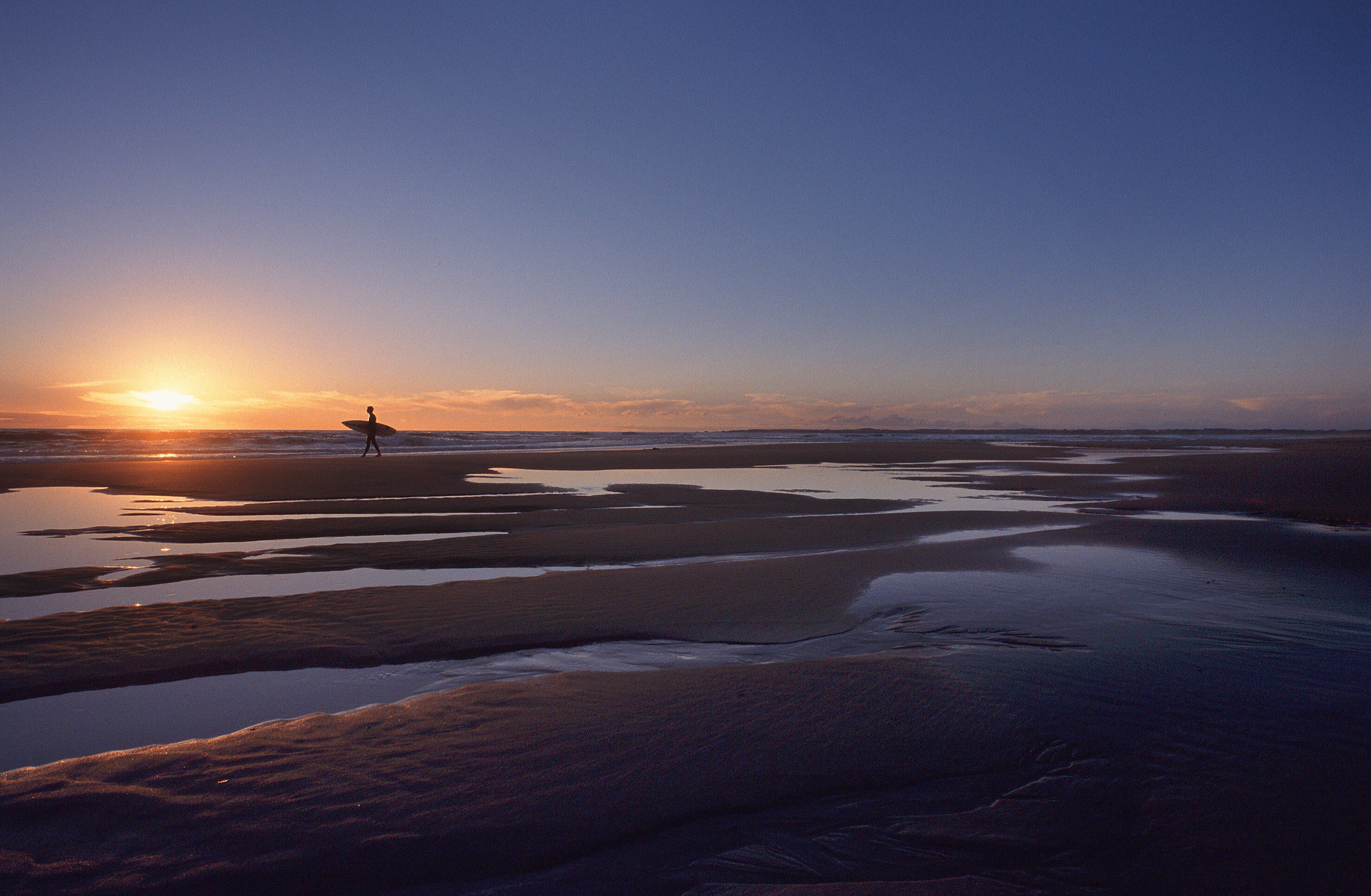 A single surfer walking across the sand at dawn at one of the best beaches in Victoria.