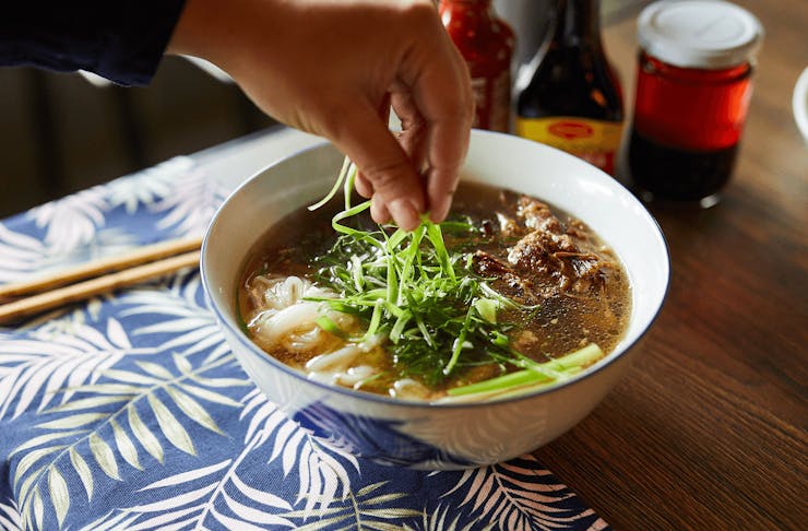 A bowl of pho with a hand dropping herbs on top.