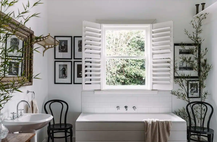 A bath sits in the centre against the wall in a bathroom, underneath a window with its blinds wide open.