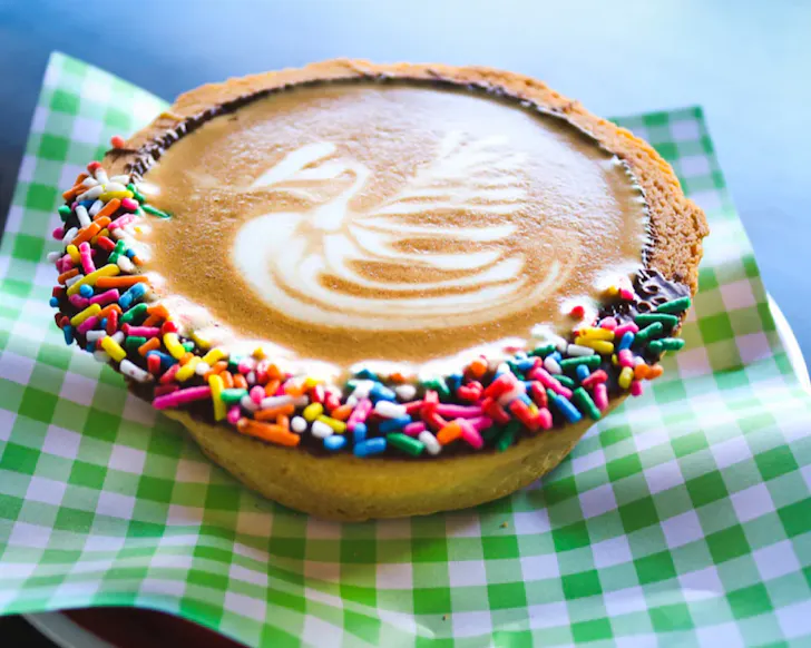 A coffee in a biscuit pie cup with a chocolate rainbow sprinkle rim.
