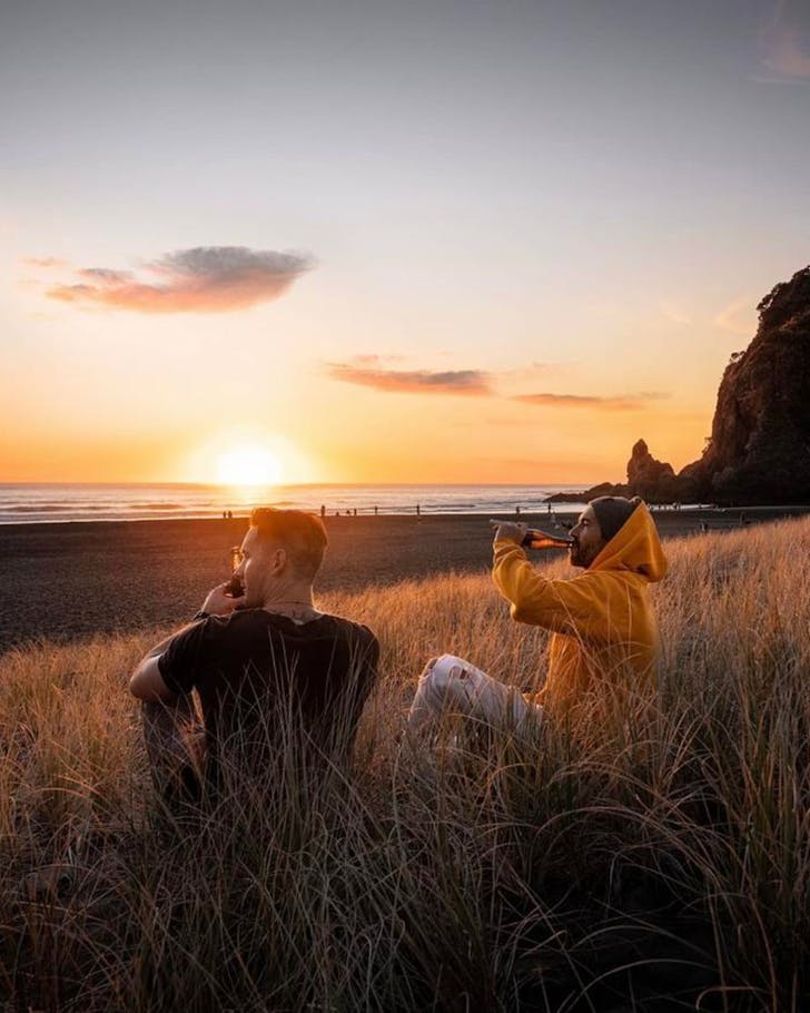 Two people enjoying beers at sunset on Piha beach. 