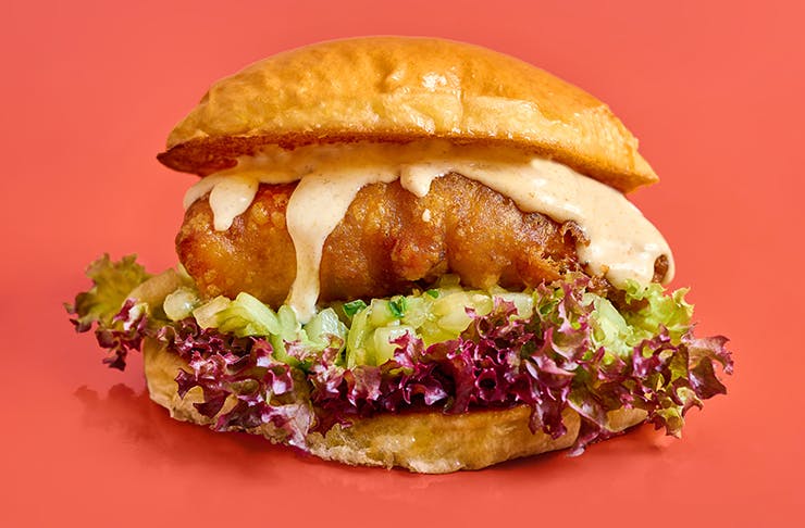 a deep fried moreton bay bug with mayo oozing down onto a bed lettuce in between a brioche bun with a red pastel background 