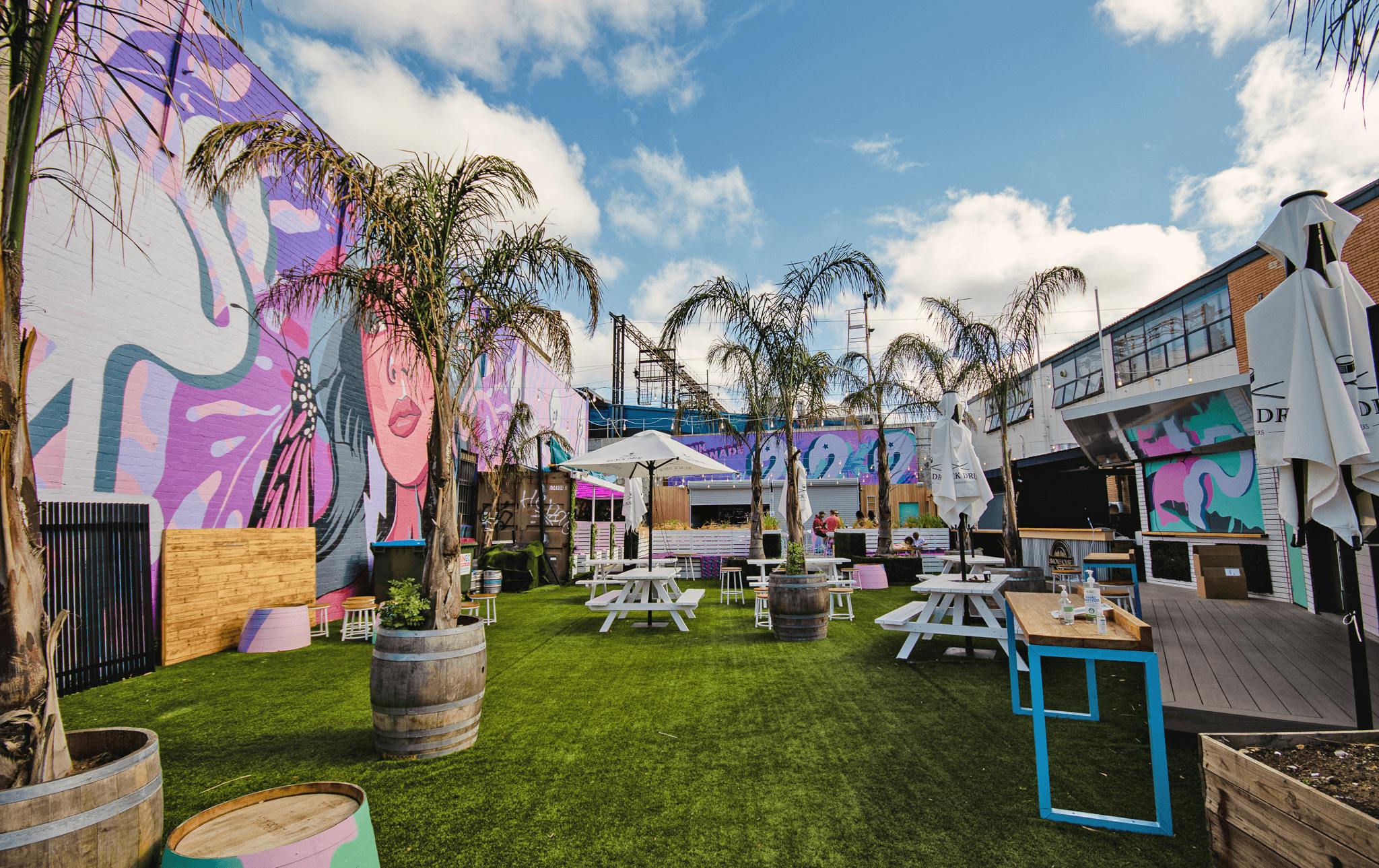 An outdoor garden bar with palm trees, lawn, and pink coloured art murals.