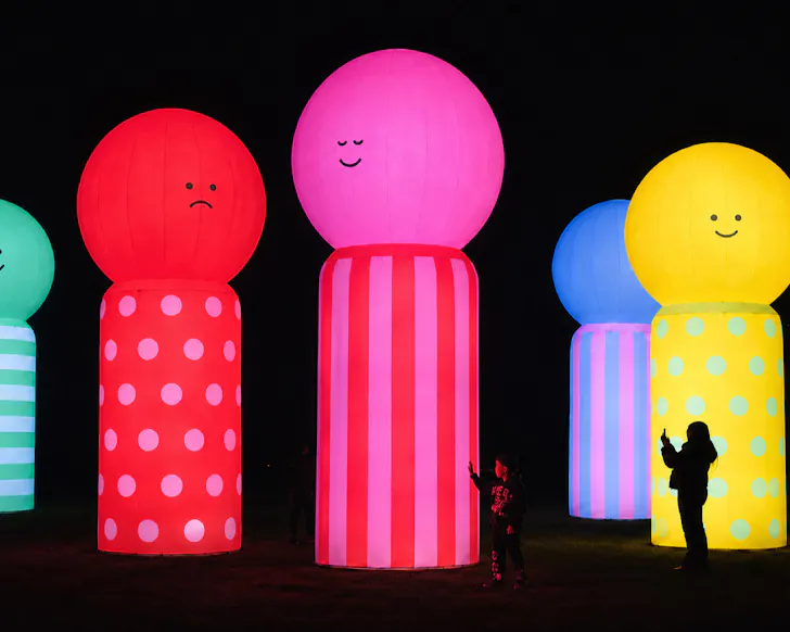 Giant inflatable lollypop people are lit against a dark night and children photograph them.