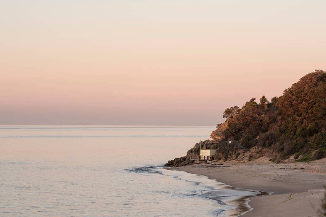 A building sits on the coastline and is soaked in a pink sunset.