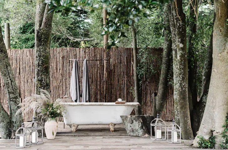 A white free-standing bath sits on a timber outdoor flooring, surrounded by trees with a wooden fence behind it.