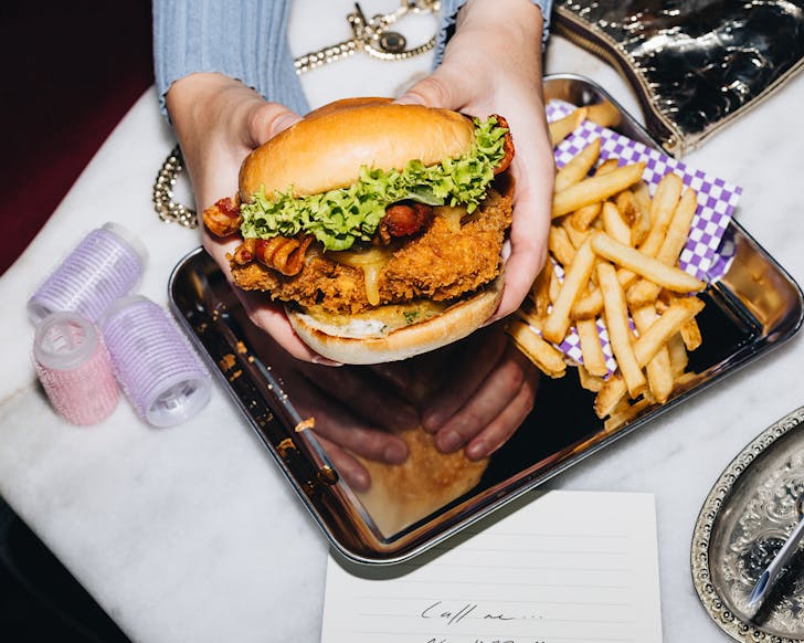 Someone holds up a burger brimming with fried chicken, bacon and curly lettuce. 