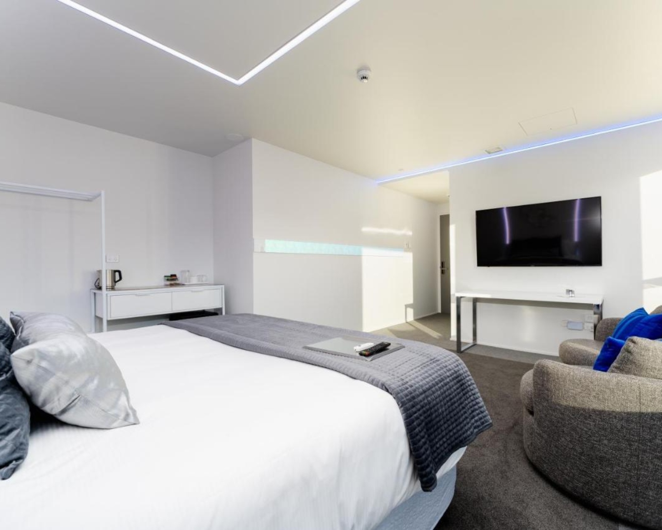 A clean neo-futuristic white apartment bedroom with a large bed, circular seats, a TV mounted on the wall and neon strip lighting on the ceiling.