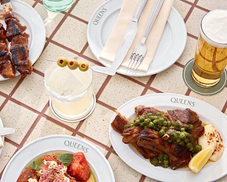 A table laden with meaty plates, a stacked tomato salad, an ice cold beer and a dirty cocktail at Queens Rooftop and Wine Shop, one of the best restaurants in Auckland. 