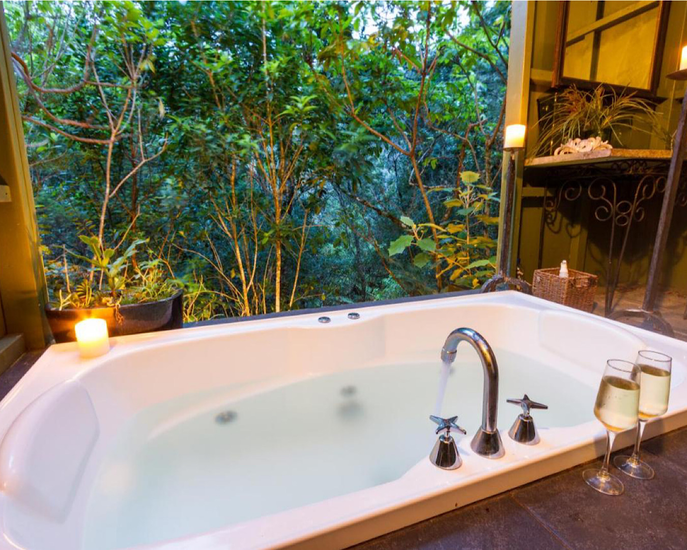 A swish bathtub slowly fills with hot water; surrounding candles burn gently and we look out to a native bush. Two glasses of white wine sit nearby. 
