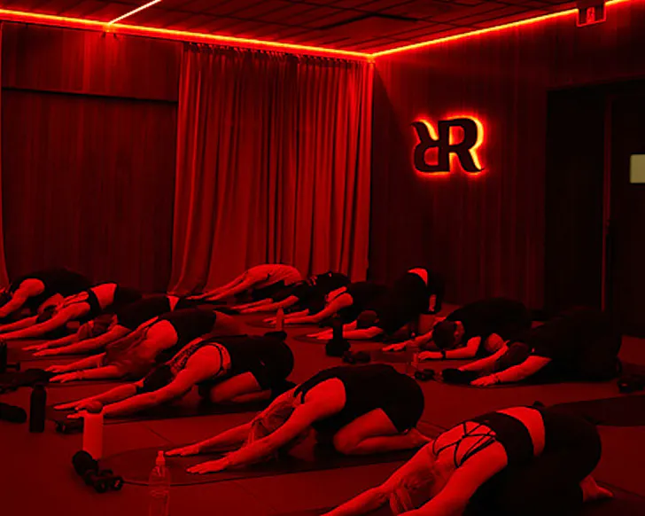 A group of people take a pause in child pose, in an infrared-heated red movement studio.