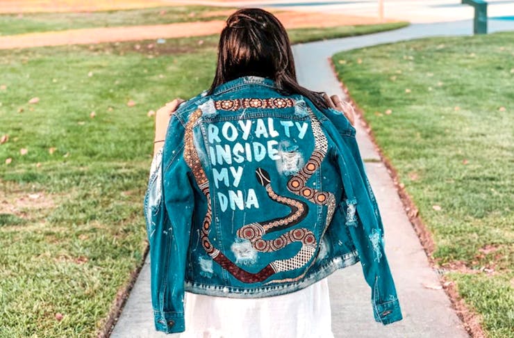 Ginny with her back to the camera wearing a jacket that reads Royalty Inside My DNA. The jacket is denim and painted with Aboriginal art.