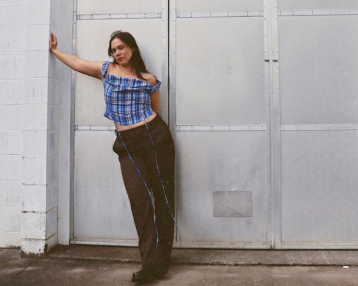 A woman wearing a blue and black chequered top, a long dark skirt and boots leans against a wall. 