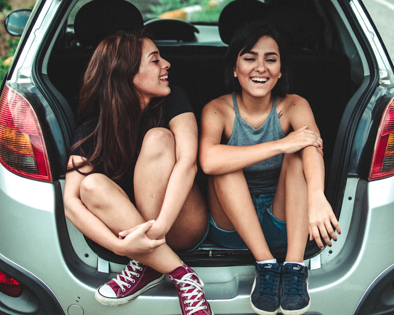 Two girls sit in the open boot of a car laughing about something. 