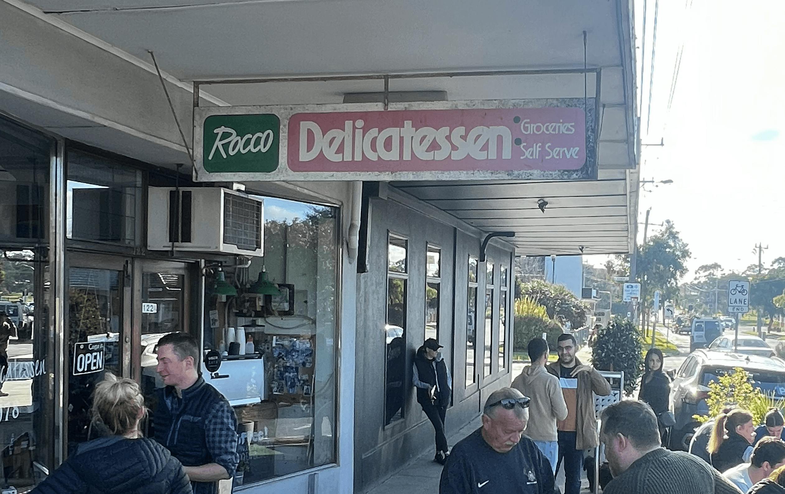 The outdoor of a deli with people standing around.