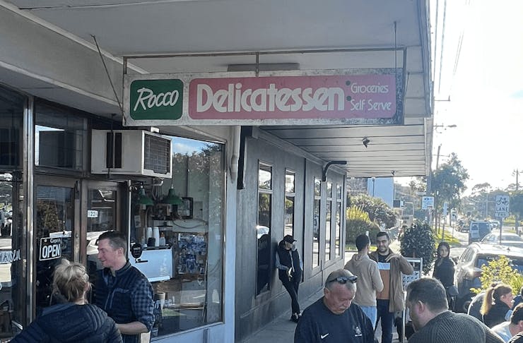 The outdoor of a deli with people standing around.