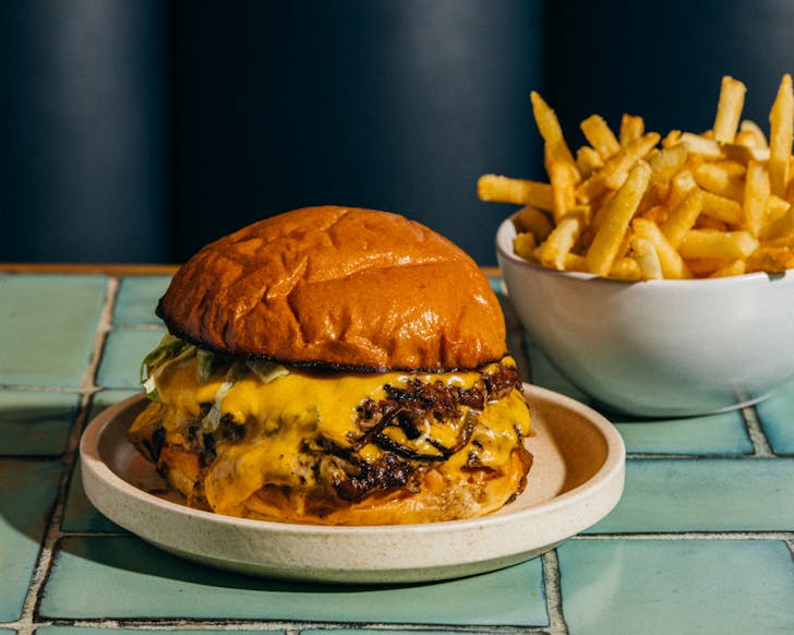 A close up image of the Rocker classic cheese burger which has a juicy paddy and melted cheese between crispy brioche buns. A small set of hot chips is in the background, also on the table.