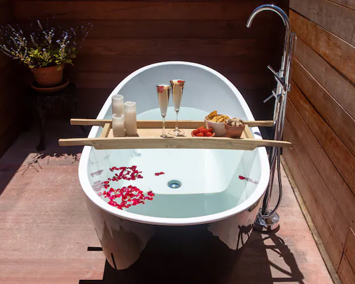 A white bathtub for two people is studded with rose petals at Rose Cottage, a beautiful romantic getaway in Auckland. A wooden ladder sits over the top with candles, champagne and nibbles. One of the best romantic getaways in auckland. 