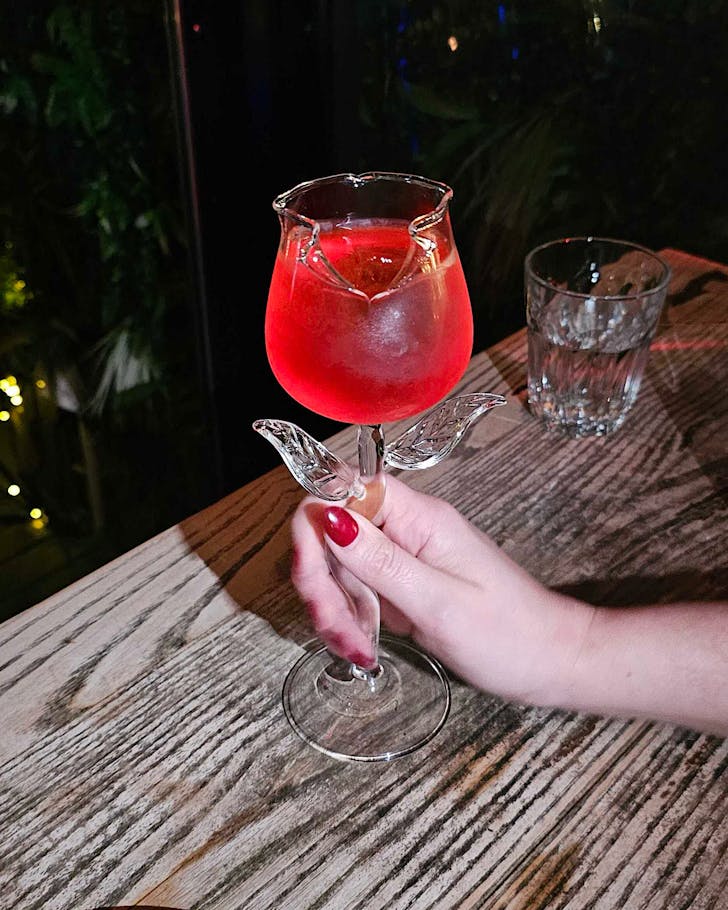 Rachel holds a rose-shaped glass of red cocktail with matching nails. 