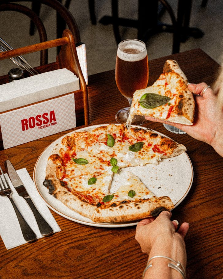 Margarita pizza on a plate atop a dark brown wooden table. Next to the pizza sits a napkin box with a pink and white checkered sticker that has ROSSA PIZZERIA BAR written on it in red type.