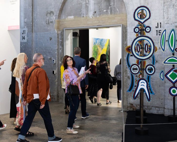A small crowd of people are walking into another room of Carriageworks, with some contemporary artworks visible on the side of the image. A woman in the middle of the small crowd has turned around to chat and discuss the artwork with a man behind her, while other people continue to walk into the next room.