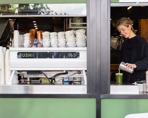 A mid shot of the outdoor window counter of Rushi, with a barista adding the final touches to an iced matcha latte, with a smile on her face. The cafe colours are sage green.