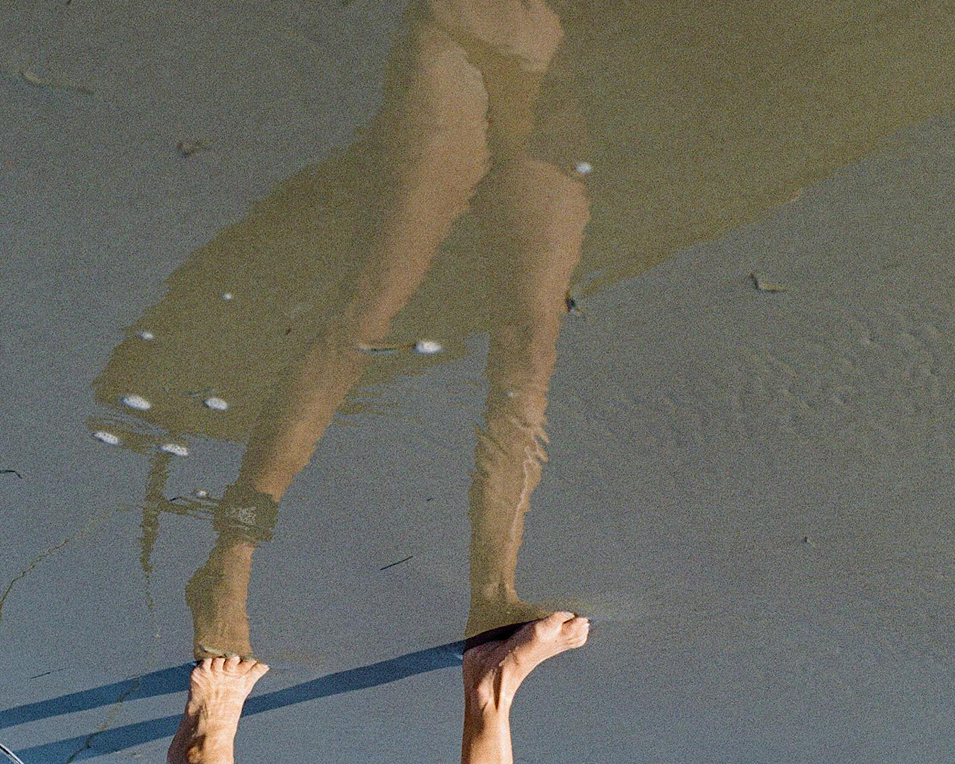 The reflection and feet of someone walking on a beach wearing a caramel-coloured bikini by Salty Sea, one of the coolest swimwear labels. 