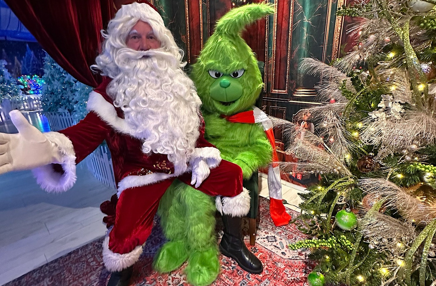 Santa sits on the Grinch's knee in Christmassy surrounds. 