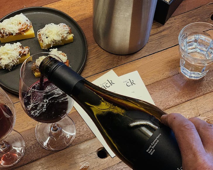 A glass of red wine is being poured while in the background some substantial snacks topped with part-melted grated cheese sit on black plates. 