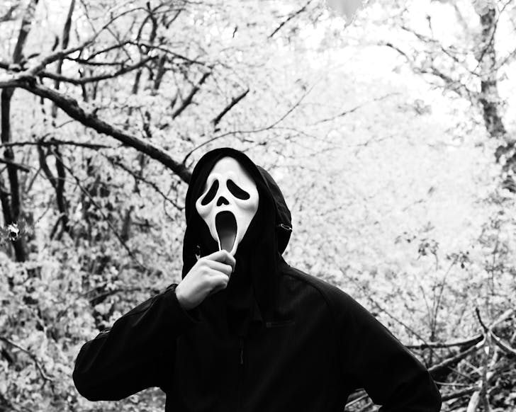 A black-and-white image of someone wearing a black cloak and Scream mask, and holding their chin while standing in a forest. 