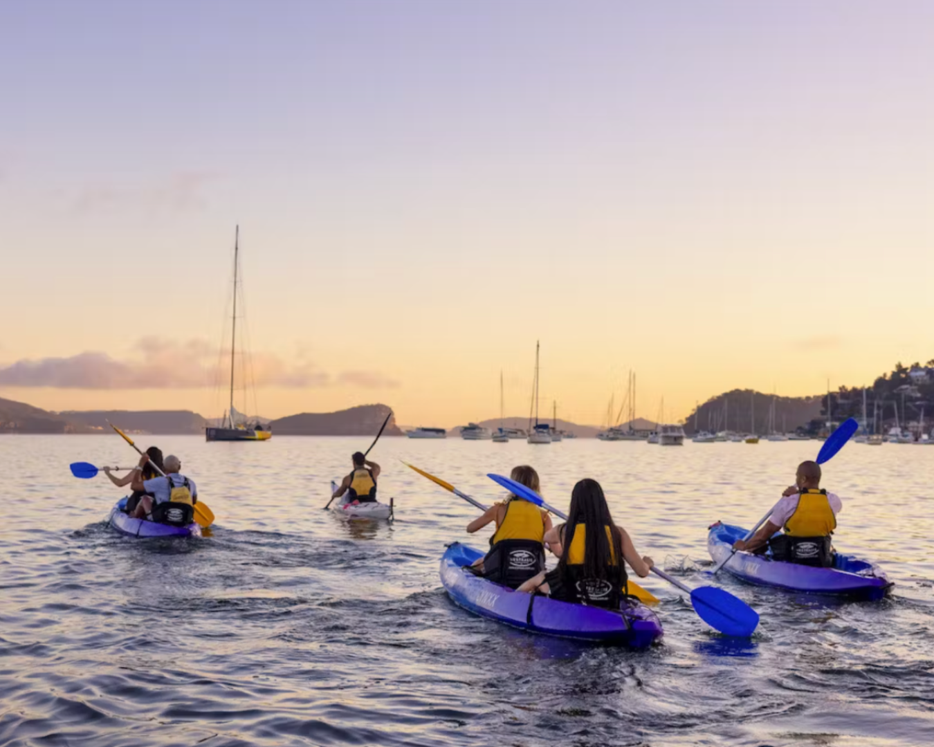 A photo following behind 4 kayaking boats paddling on the water. They are facing away from the camera and paddling towards the sunset.