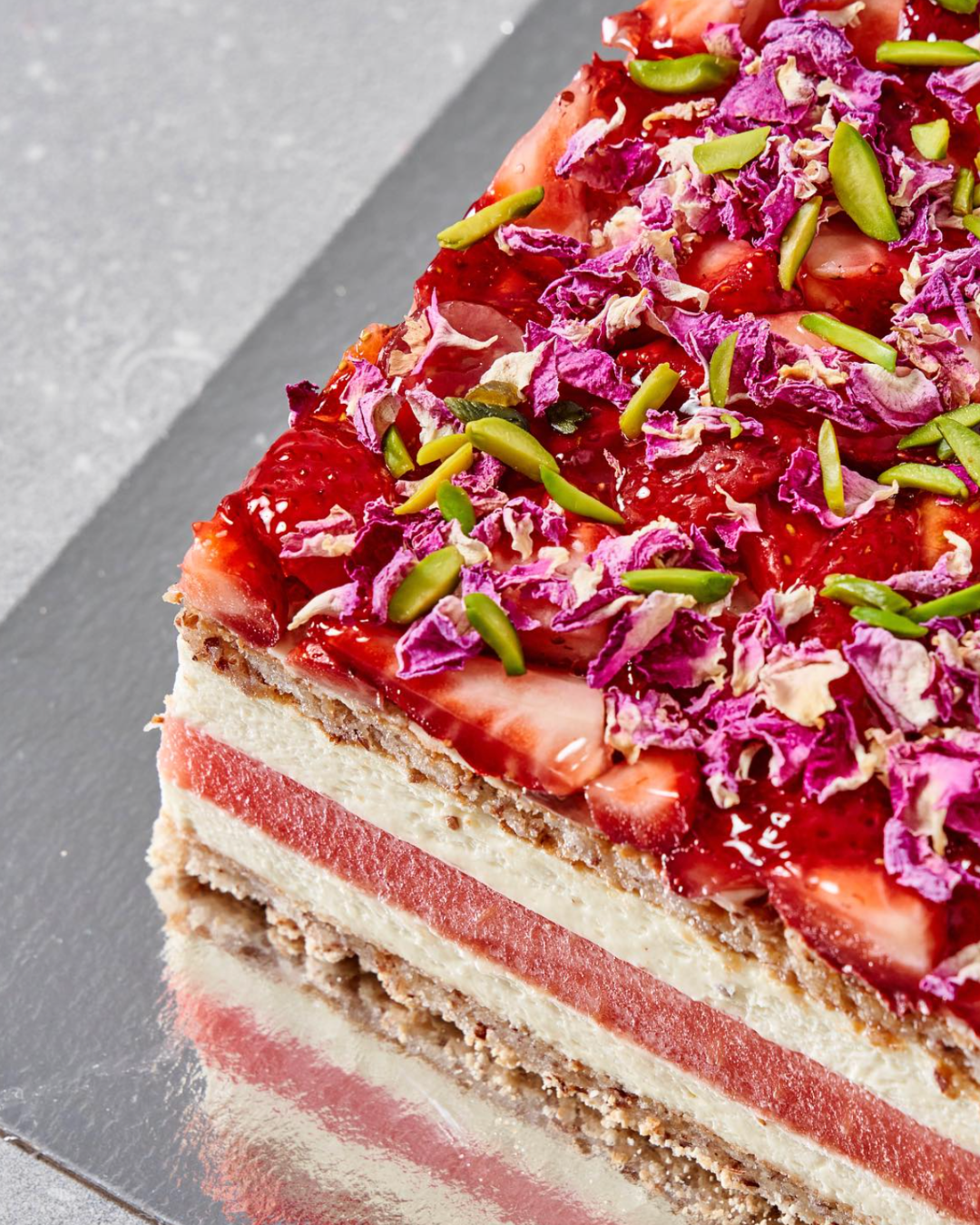 A close up picture of the Strawberry Watermelon Cake. Topped with a thick layer of strawberries and rose petals, the side of this cake reveals the layering of fresh watermelon in the middle, cream on either side, and sandwiched on the outer layers with almond dacquoise.  