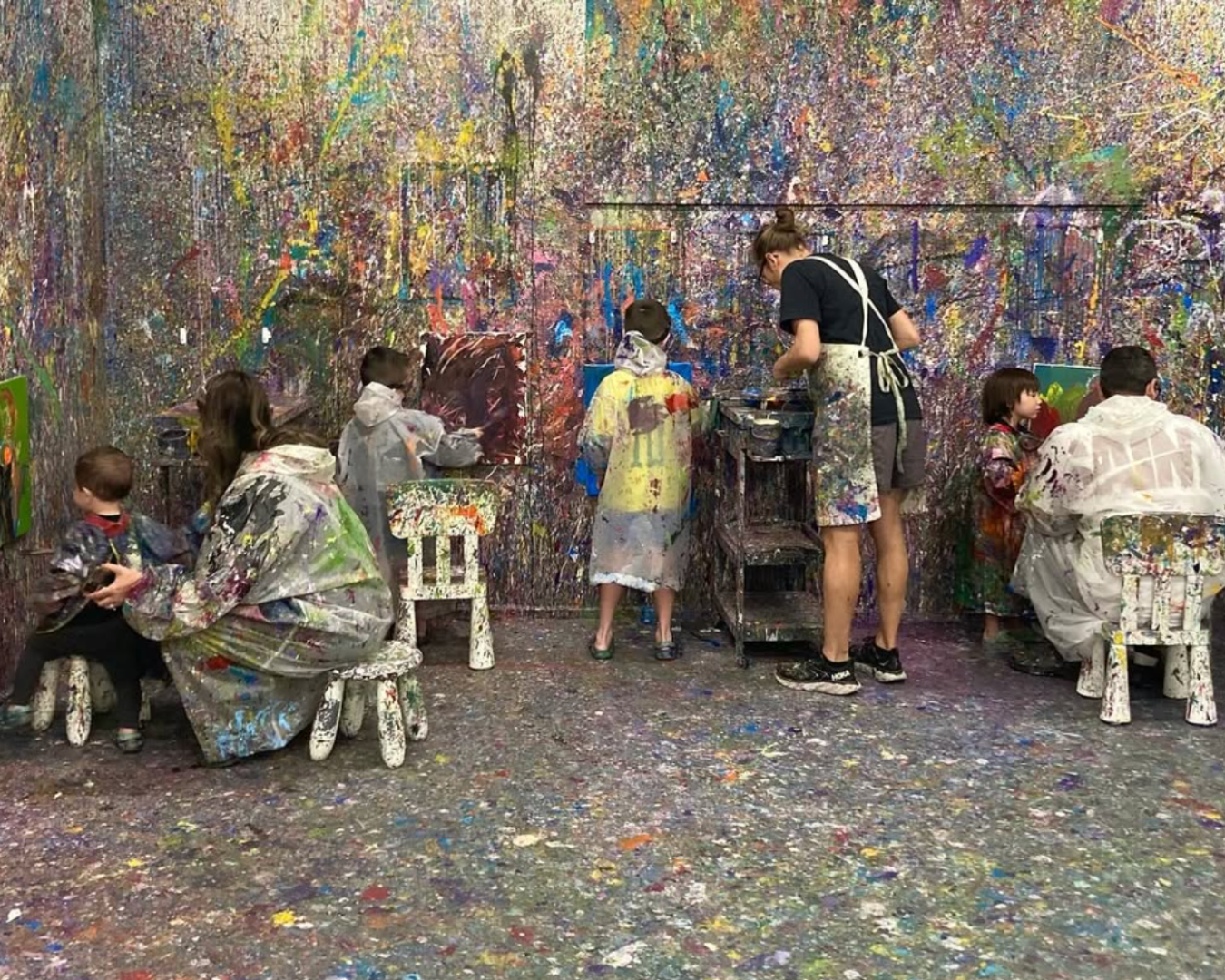 A wide shot of a splatter paint room with walls covered in a colourful mess of paints. A few children and adults stand in front of their canvases on the walls, with all of them wearing paint coats and goggles.