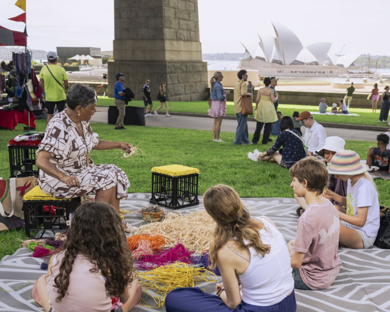 A wide shot of a woman sitting on a picnic mat with children sitting around her as she is teaching them how to weave. There are colourful weaving threads in the middle of the picnic blanket. In the background, there are some other stalls and a blurred view of the Sydney Opera House across the water.