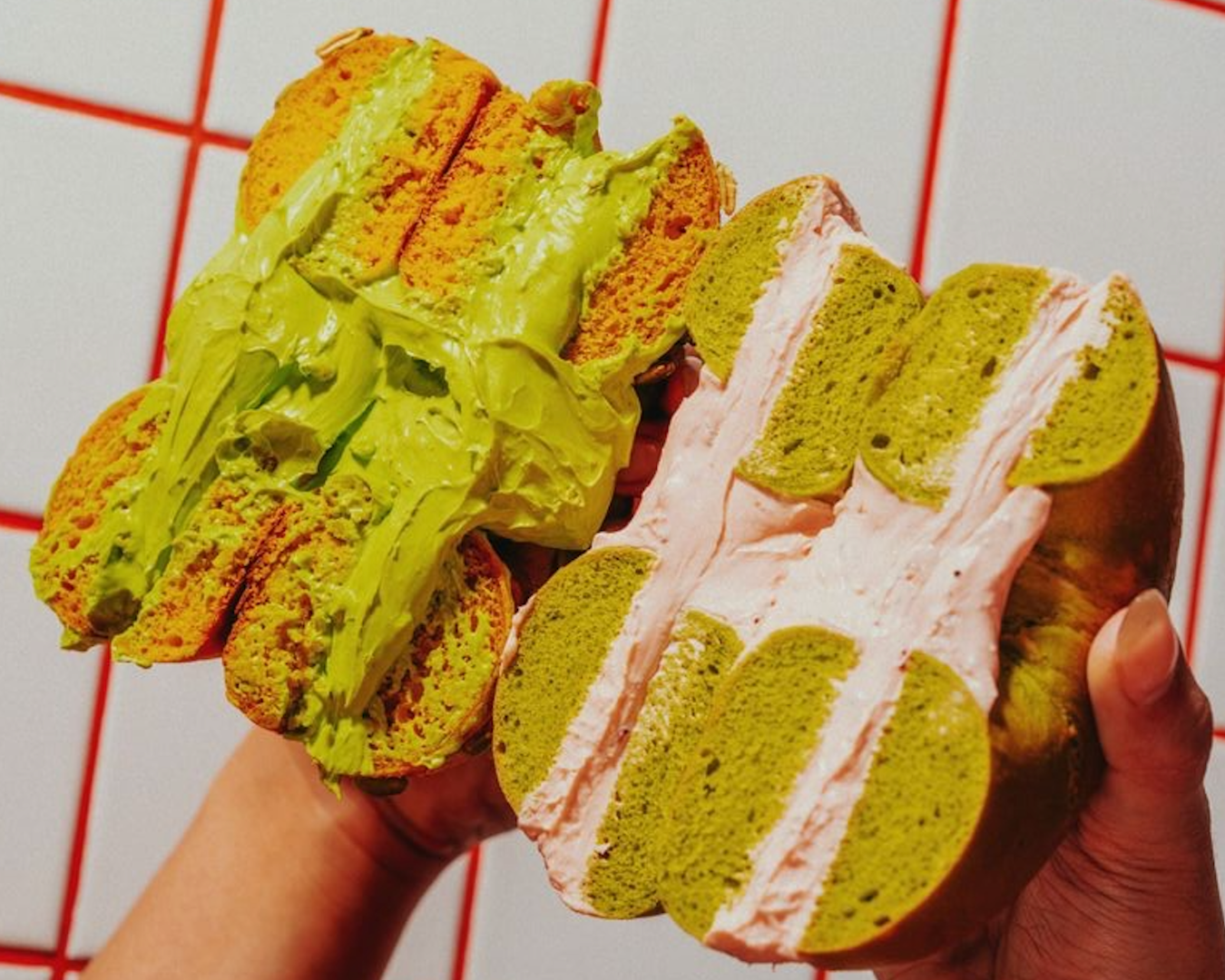 A close up image of two bagels cut in half with the inside filling showing. One has thick matcha green cream oozing out, and the other has a light pink cheese cream and the bagel itself is green matcha coloured.