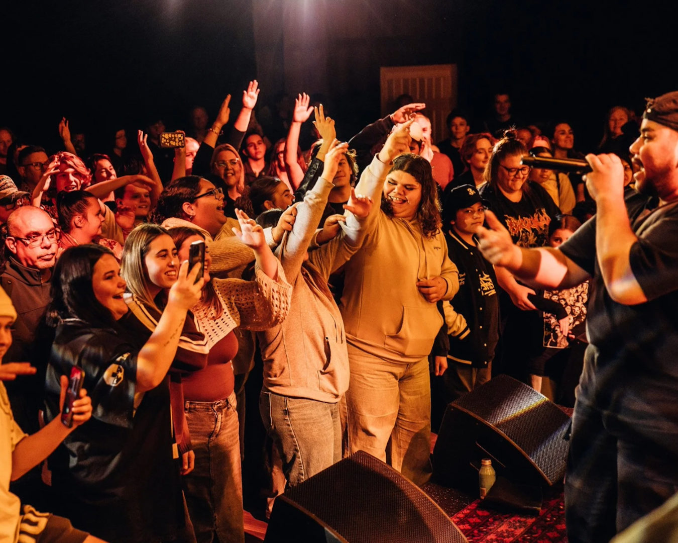 A wide shot of a crowd of people dancing in a dark room, looking at the performer who is singing to the far right of the image. The image is taken from the stage next to the performer.