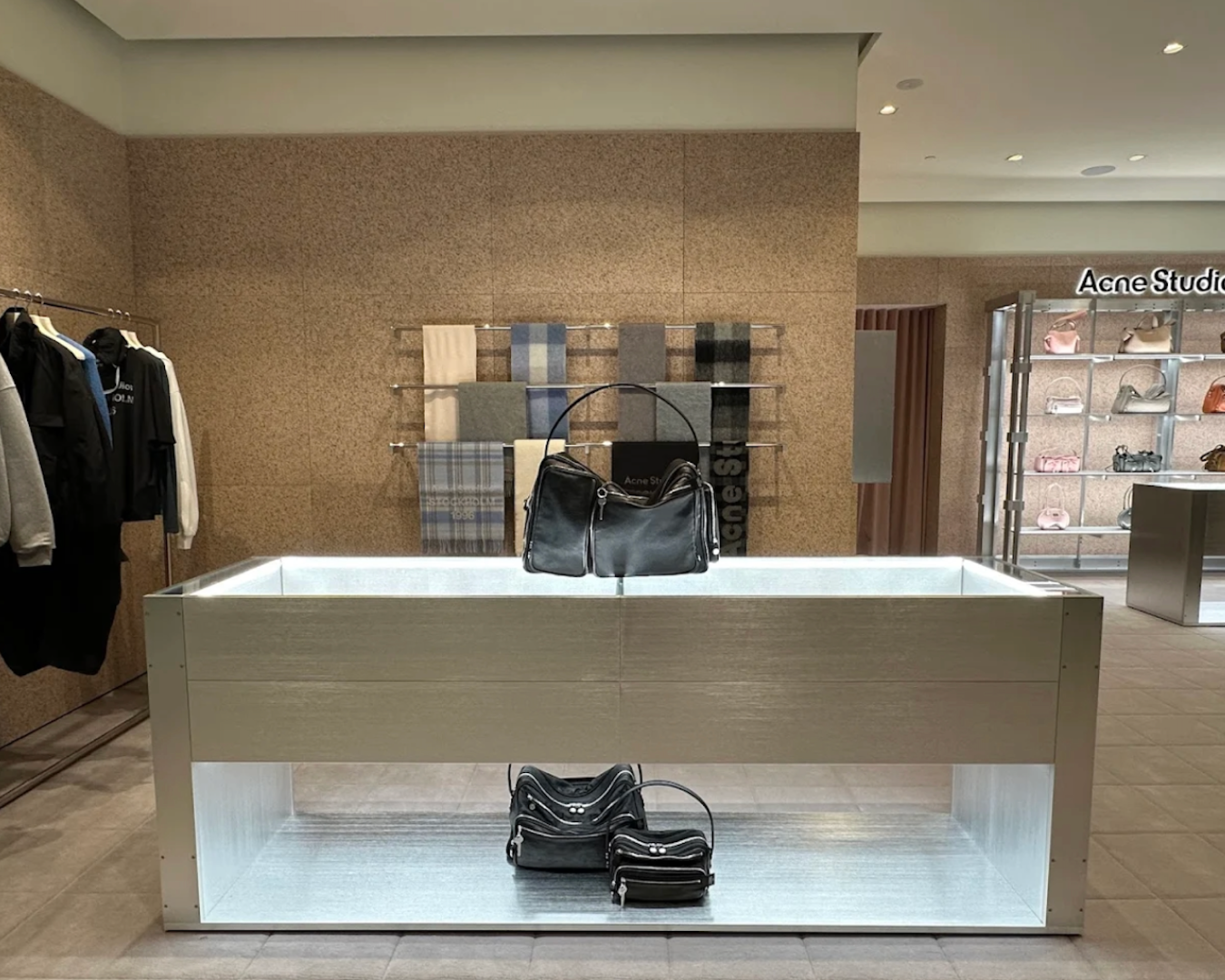 A wide shot of inside the Acne Studio store in Westfield focusing on a bag sitting on a display table. Around the room there are scarves on display, clothing on hangers, and a sign that says Acne Studios. 