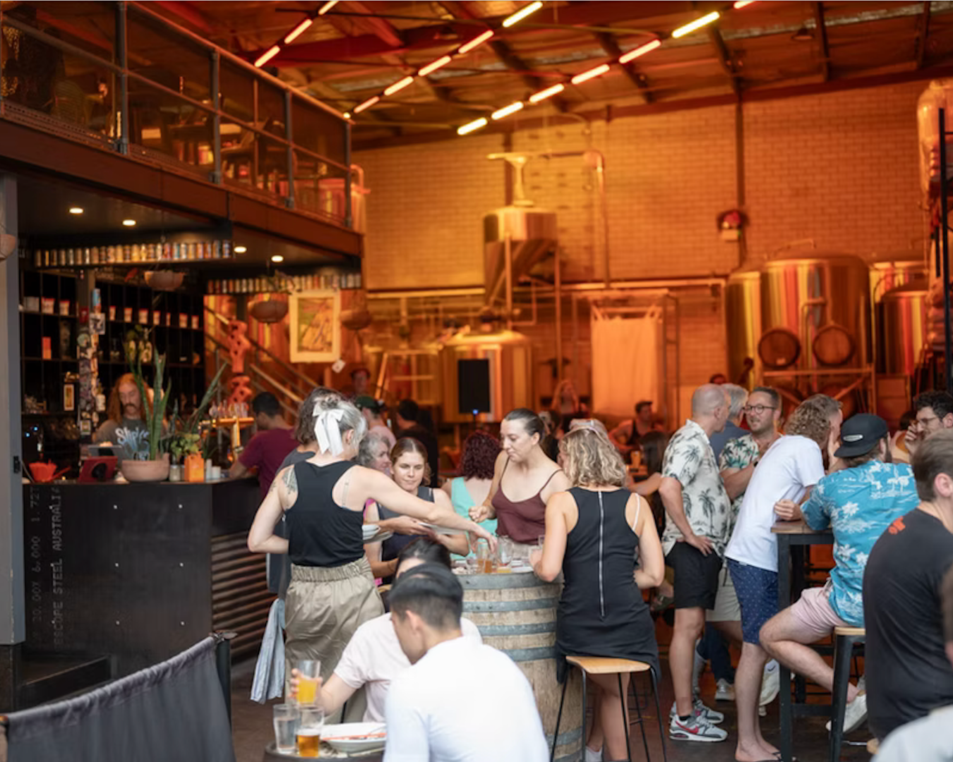 A wide shot of the interior of Yulli's Brews showing many full tables of people chatting and drinking, a bar to the left of the image and some large brewery machines in the background.