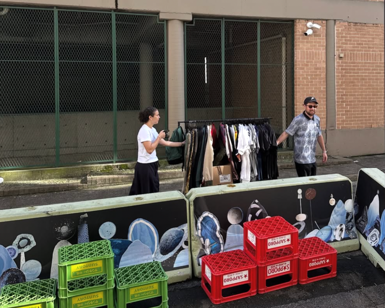 A mid shot of the exterior of Poketto Market where two people are moving a rack of clothing on wheels, seemingly in the process of putting it inside of the cafe and setting up the market stalls.