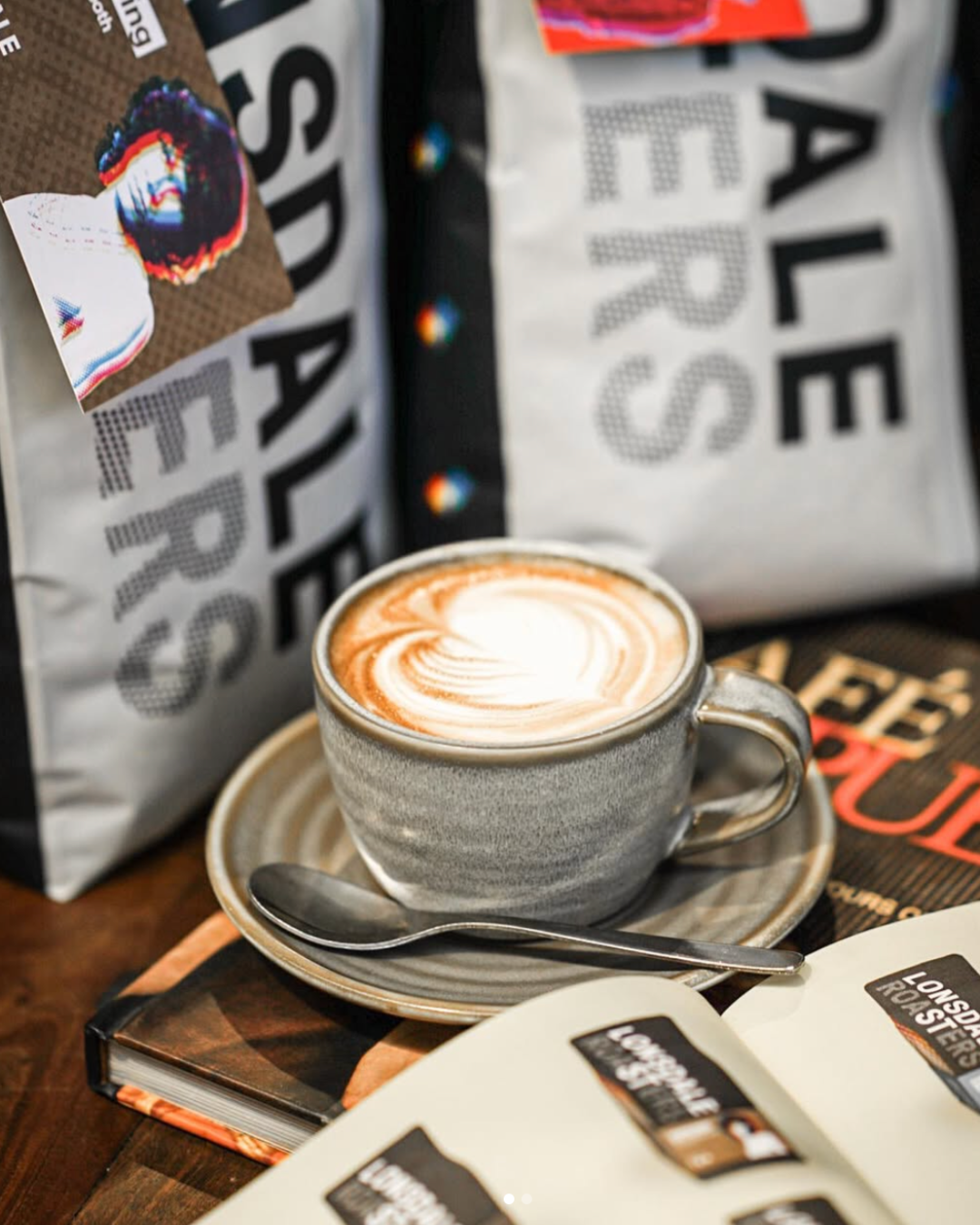 A close up image of a coffee in a ceramic cup and saucer balanced on a table that is surrounded by Lonsdale Street Roasters bean and merchandise.