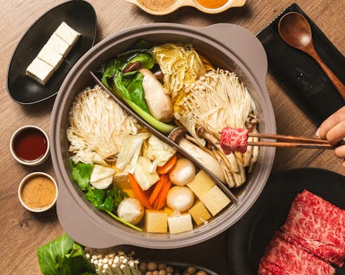An above, birds-eye view of the yin-yang hot pot bowls full of soup and ingredients. A person's hand is using chopsticks to cook a piece of raw meat in the hot pot.