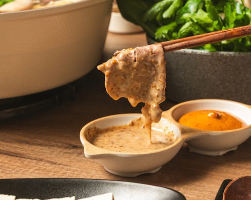 A close-up picture of a piece of cooked meat being held by chopsticks. The meat has just been dipped into a ramekin full of sauce, and the meat is now covered in this sauce.