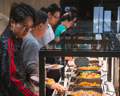 A line of people are in front of the self-serve buffet section. They are each serving themselves a selection of foods including rice, vegetables and meats.